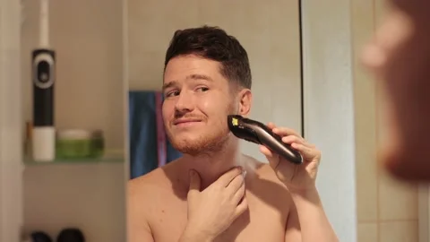 A cheerful Man equalizes his beard with a trimmer in the bathroom, looking in Stock Footage 240056886