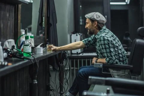 Cheerful man in flat cap drinking alcohol while being in barber shop Stock Photos