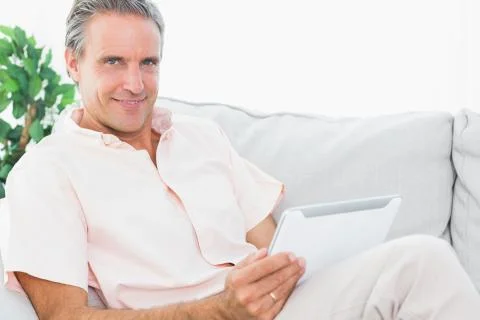Cheerful man on his couch using tablet pc looking at camera 스톡 사진