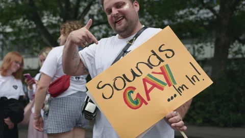 A cheerful man points at a handmade sign reading Sounds GAY I'm In with rainbow Stock Footage 346238818