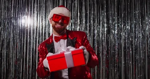 Cheerful man in Santa hat dancing with gift box against shiny backdrop at Stock Footage 320440812