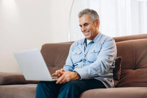 Cheerful man sitting on sofa, using laptop at home in cozy living room. Casual Stock Photos