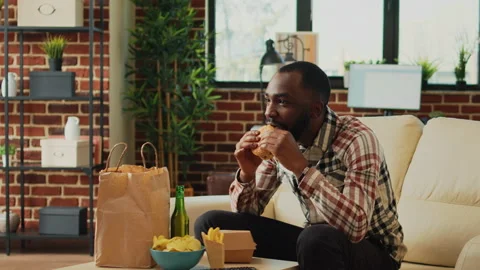 Cheerful man taking bite of hamburger in living room Stock Footage 231079536