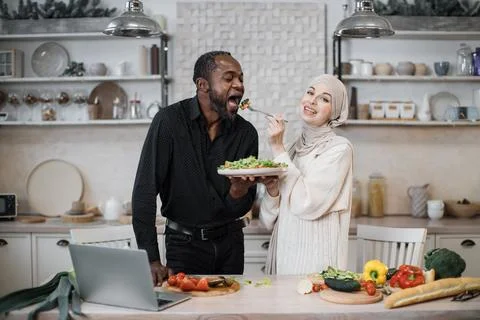 Cheerful married multinational couple using laptop while cooking healthy food in 스톡 사진