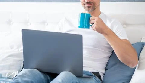 Cheerful mature man using computer in bed with coffee Foto stock