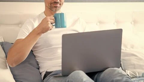 Cheerful mature man using computer in bed with coffee Foto stock