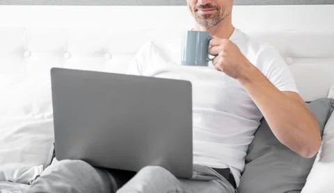 Cheerful mature man using computer in bed with coffee 写真素材