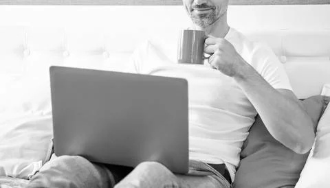 Cheerful mature man using computer in bed with coffee Foto stock