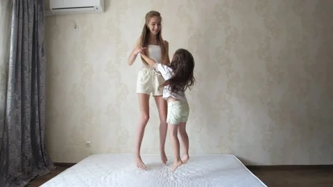A cheerful mom plays with a cute active little daughter, jumping on the mattress Stock Footage 210455511