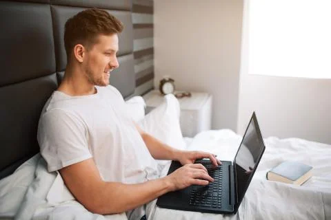 Cheerful nice young man work in bed early moring. Guy hold laptop and type on 写真素材