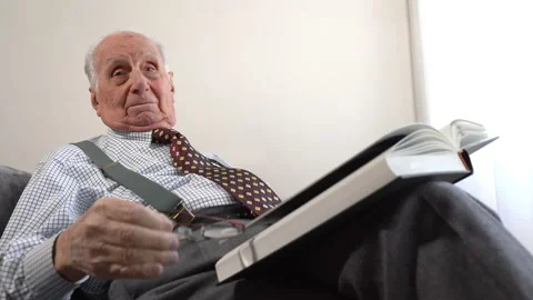 Cheerful old man sitting on the sofa in living room, holding eyeglasses in hands Stock Footage 230039949