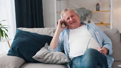 Cheerful old man talking by phone while sitting on the sofa at home Stock Footage 301779907