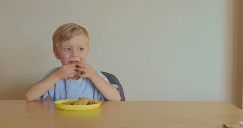 A cheerful, satisfied boy sits at a table, dancing slightly to music and eating Stockbeeldmateriaal 324819613