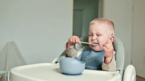 Cheerful toddler boy eats milk porridge with a spoon while sitting in a high Stock Footage 315535956