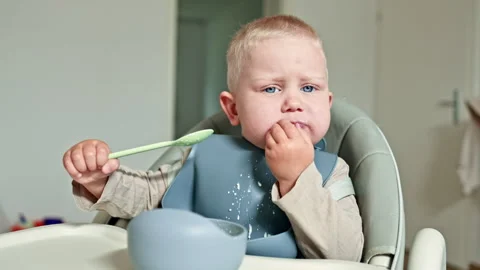 Cheerful toddler boy eats milk porridge with a spoon while sitting in a high Stock Footage 319935447