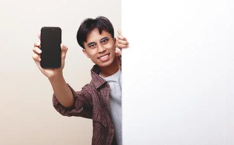 Cheerful young Asian man peeking from behind blank white board while showin.. Stock Photos
