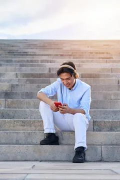 Cheerful young Asian man using mobile phone outdoors, while listening music Stock Photos