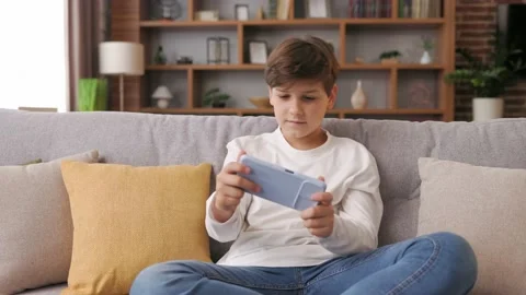 Cheerful young boy playing video game on smartphone sitting on sofa at home Stock Footage 203796261