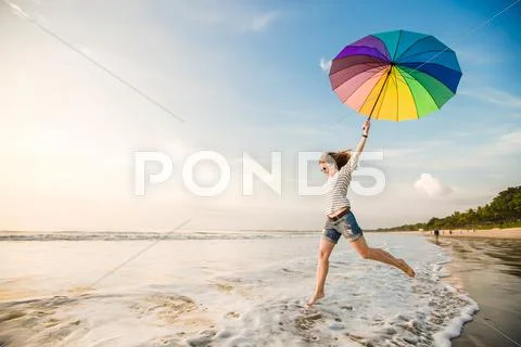 Cheerful Young Girl With Rainbow Umbrella Having Fun On The Beach Before Sunset
