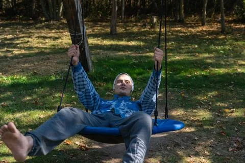 A cheerful young guy riding a swing in the village. A guy in a shirt and a scarf Stock Photos