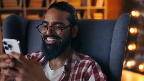 Cheerful young Indian man chatting on phone at home library, smiling at screen Stock Footage 313282533