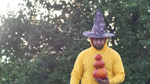 A cheerful young man in a black witch's hat holds a pyramid of pumpkins in his Stock Footage 213899508