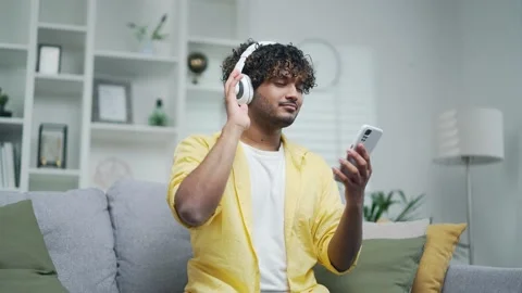 Cheerful young man in a bright yellow shirt listens to music in headphones using Stock Footage 235505075