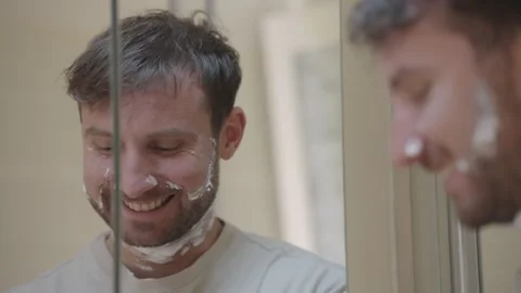 Cheerful young man making funny faces while applying shaving cream in front of a 스톡 동영상 335373872
