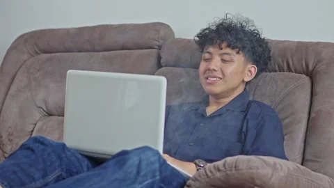 Cheerful Young man relaxing using laptop while exhaling vape clouds on couch Stock Footage 332390815