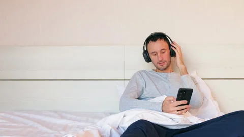 Cheerful young man sitting on bed in bedroom and enjoying music with headphones. Stock Footage 166327907
