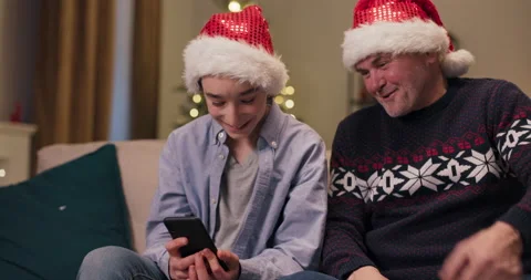 Cheerful young man sitting on couch with grandfather and holding phone. He shows Stock Footage 210608325