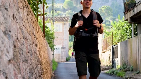 Cheerful young man walking with a backpack through a village with green mou.. Stock Footage 294178122