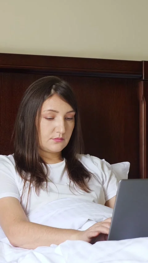 Cheerful young woman using her laptop while comfortably lying in bed, enjoying a Stock Footage 342912575