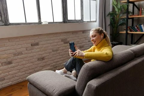 Cheerful young woman using mobile phone while sitting on a sofa at home. Stock Photos