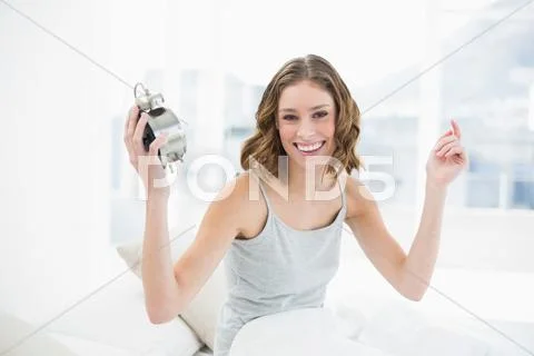 Cheering young woman holding an alarm clock sitting on her bed Stock Photos