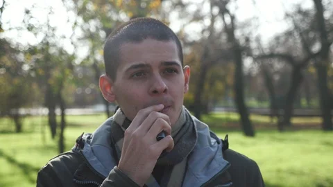 Cheery young man vaping an e-cigarette in a park in autumn in slow motion Stock Footage 120419063