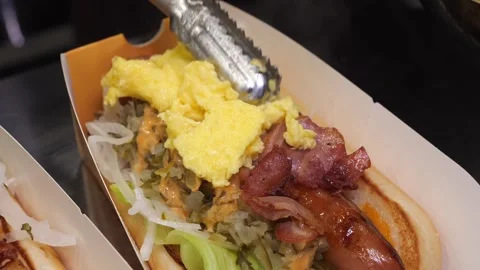 Chef adding scrambled eggs topping on gourmet hot dog with bacon and sausage Stock Footage 324711093