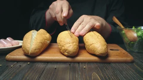 Chef in black attire skillfully slices open bread rolls for hot dogs on a wooden Stock Footage 337027118