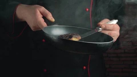 Chef in black coat flips cooked meat in frying pan, showcasing culinary skills Stock Footage 337904462