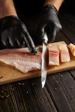 A chef with black gloves cuts a fish fillet on a wooden board. The fillet is  Stock Photos