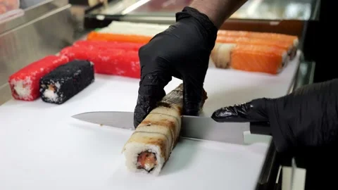 A chef in black latex gloves prepares sushi, he slices sushi with a large knife. Stock Footage 322215468