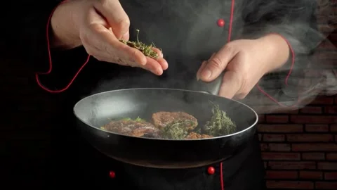 Chef in black uniform sprinkles dry herbs over sizzling meat in a frying pan, Video stock 337512231