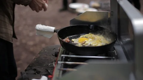 Chef cooks eggs and bacon in a pot over a camping stove Stock Footage 91276444