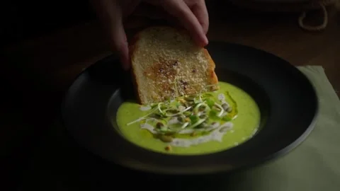 A chef dips crispy toasted bread into creamy zucchini soup garnished with Stock Footage 342543363