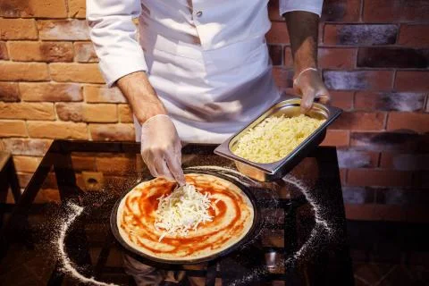 Chef in gloves prepares pizza, adding cheese on it. Pizza cooking process. Stock Photos