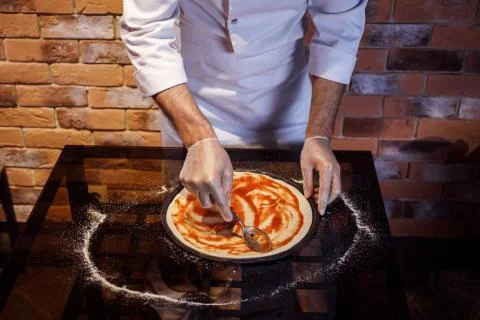 Chef in gloves prepares pizza, adding tomato sauce on it. Pizza cooking process. Stock Photos