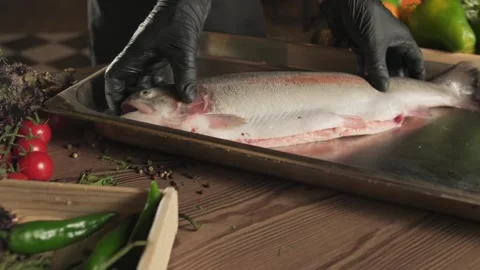 Chef in gloves salting fish on kitchen table among herbs, tomatoes and Stock Footage 270723482