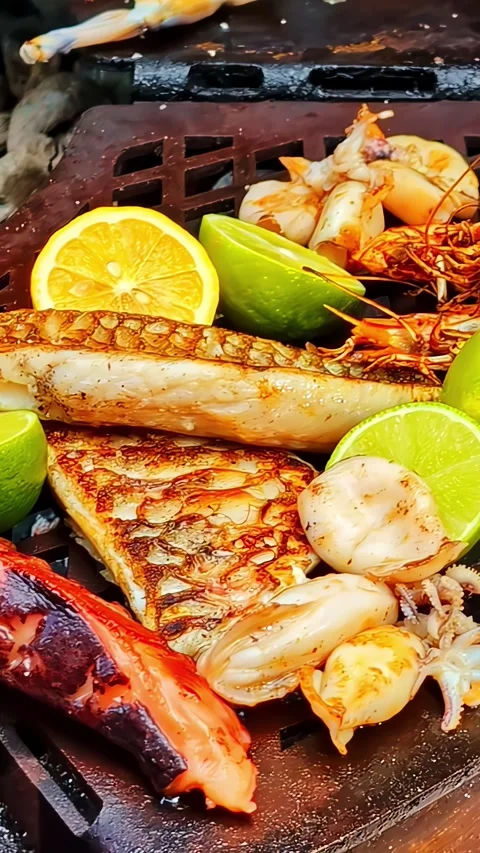 Chef grilling frog legs and seafood on a hot griddle, with tongs, prawns, lime Video stock 336242969