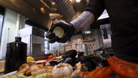 Chef hand in black chef rubber gloves squeezing lime juice in fried mussels 스톡 동영상 105272075