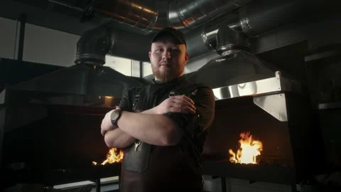 Chef with leather apron stands with folded hands near burning barbecues Stock Footage 315230130